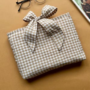 Può includere: Una pochette a quadretti marrone e bianca con fiocco abbinato, occhiali rotondi neri e un libro con testo turco. La pochette è rettangolare e ha un grande fiocco sopra. Il libro è aperto.