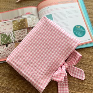 May include: A pink and white gingham fabric pouch with a matching bow. The pouch is open, revealing the interior. An open book and a pair of glasses are in the background. The book has text in German.