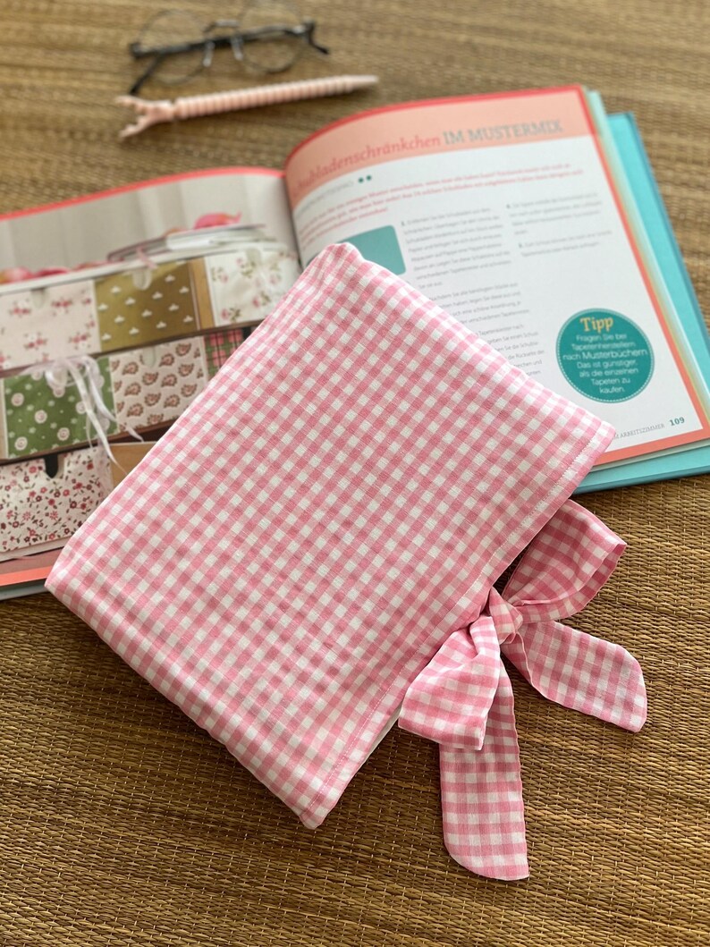 May include: A pink and white gingham fabric pouch with a matching bow. The pouch is open, revealing the interior. An open book and a pair of glasses are in the background. The book has text in German.