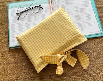 以下が含まれることがあります： 黄色と白のギンガムチェックのペンケース。リボン結びで、開いた本の上に置かれています。本にはドイツ語のテキストと丸いメガネが写っています。