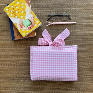 May include: A stack of books, including one with a yellow cover and the title "Kendime Ait G&ouml;r&uuml;nmez Bir İşaret". Also, a pair of round eyeglasses, a pink pen, and a pink and white gingham fabric pouch with a bow.