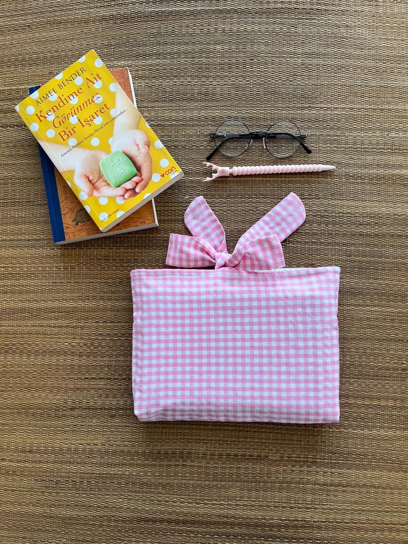 May include: A stack of books, including one with a yellow cover and the title "Kendime Ait G&ouml;r&uuml;nmez Bir İşaret". Also, a pair of round eyeglasses, a pink pen, and a pink and white gingham fabric pouch with a bow.