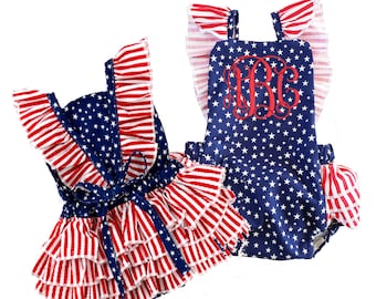 May include: Two baby outfits in red, white, and blue with a star pattern. One outfit is a ruffled dress with a bow and the other is a one-piece with a monogram.