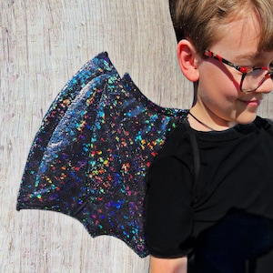 Puede incluir: Un niño con una camisa negra y alas de dragón iridiscentes. Las alas están hechas de un material brillante y tienen una base negra con purpurina arcoíris.