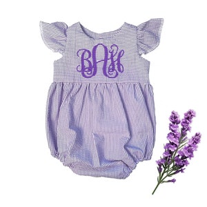 May include: A purple and white striped baby romper with flutter sleeves and a bubble bottom. The romper features a purple monogram on the chest. A sprig of lavender is next to the romper.