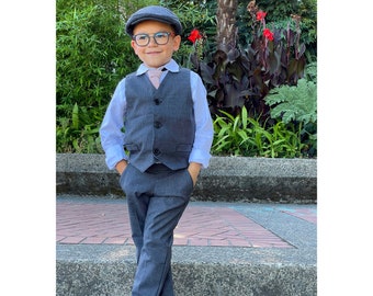 Op de afbeelding: Een kind draagt een grijs pak bestaande uit een vest en een broek, met een wit overhemd en een roze stropdas. De outfit wordt gecompleteerd met een grijze pet, een bril en bruine schoenen. De achtergrond toont groen en een bakstenen pad.