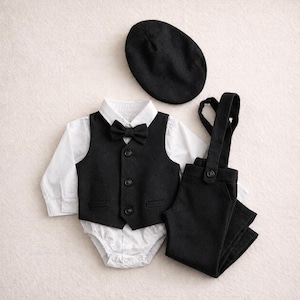 May include: A formal baby outfit set, including a black vest, pants with suspenders, a white shirt, a black bow tie, and a black cap. The vest has three buttons. The outfit is designed for a baby or toddler.