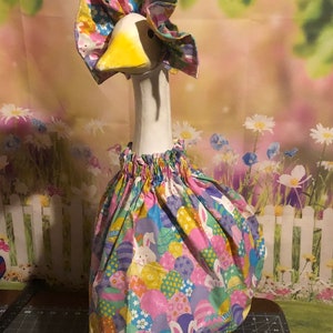 May include: A white ceramic goose figurine wearing a pink and green Easter-themed dress with bunnies and eggs. The goose has a yellow beak and a ruffled pink collar.