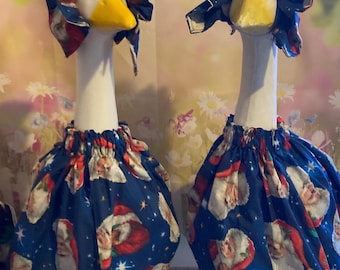 May include: Two goose figurines dressed in festive Christmas attire. Each goose has a white neck and a yellow beak. They wear blue dresses and bonnets patterned with Santa Claus faces and stars.