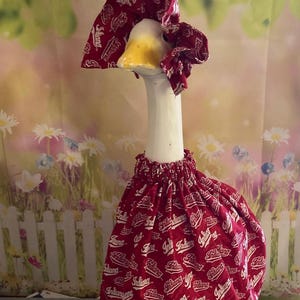 May include: A decorative goose figurine wearing a red dress and matching head covering. The fabric features a repeating white text pattern with the word "Indiana". The goose has a white neck and head with a yellow beak.