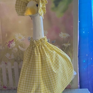 May include: A decorative goose figurine wearing a yellow and white checkered dress and matching bonnet. The goose has a white neck, yellow beak, and is standing on a green base. The background features a floral design.
