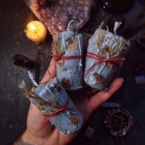 Puede incluir: Tres velas azules con hierbas secas y cordel rojo. Las velas tienen un aspecto rústico, con texturas visibles y elementos incrustados. Una vela encendida y cristales en el fondo sugieren un tema espiritual o decorativo.