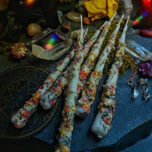 May include: A collection of white candles with dried flowers and herbs embedded in the wax. The candles are arranged on a dark surface with a gold circular design.