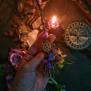 Op de afbeelding: Een kleine, handgemaakte bezem met een pentagram-hanger, versierd met gedroogde bloemen en vastgebonden met een geel lint. De bezem wordt in een hand gehouden tegen een donkere achtergrond met een brandende kaars en andere magische voorwerpen.