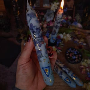 May include: Light blue candles with embedded purple flowers and gold flecks. Each candle has a blue triangle design with a gold center. The candles are lit, with flames at the top. The candles are part of a set.