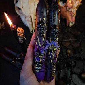 Könnte beinhalten: Zwei schwarze Kerzen mit goldenen Akzenten und Totenkopfdesigns sind mit lila und schwarzem Garn zusammengebunden. Getrocknete lila Blumen sind in das Garn eingewebt.