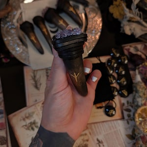 Pode incluir: Uma varinha marrom escura com uma runa dourada, encimada por um aglomerado de cristais roxos. A varinha é segurada em uma mão com uma tatuagem. Outras varinhas estão em uma bandeja de prata.