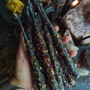 May include: A collection of dark blue taper candles, adorned with dried flowers and herbs. The candles are held in a hand, with a lit candle in the background. The candles are approximately 8 inches long.