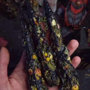 May include: Three black beeswax candles with dried herbs and flowers embedded in the wax. The candles are held in a hand.