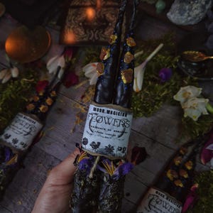 May include: Two black candles with gold and blue embellishments are wrapped in a brown paper label that reads "Moon Magician Flowers Fragrance of Memories Ritual Candles Full Moon". The candles are surrounded by dried flowers and herbs.
