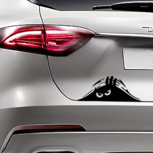 May include: A black and white car decal of a monster peeking out from behind a wall with one eye visible. The decal is applied to the back of a silver car.