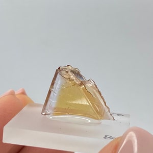 Può includere: Un cristallo trasparente, sfaccettato, di colore marrone chiaro con una leggera sfumatura rosata. Il cristallo è posto su un espositore quadrato in vetro bianco.