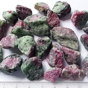 May include: A collection of rough, green and pink ruby zoisite gemstones. The stones are various sizes and shapes, with some showing a more pronounced green colour and others a more pronounced pink colour.
