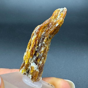 Op de afbeelding: Een ruw, langwerpig mineraal exemplaar met een vezelige, gelaagde structuur in een kleurenpalet van diep amber, goudgeel en doorschijnend wit. Het kristal heeft een grillige, onregelmatige oppervlaktestructuur en is bevestigd op een vierkante voet.