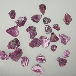 Könnte beinhalten: Eine Sammlung von rosa Rubinkristallen, einige mit Streifen, auf einer weißen Oberfläche.