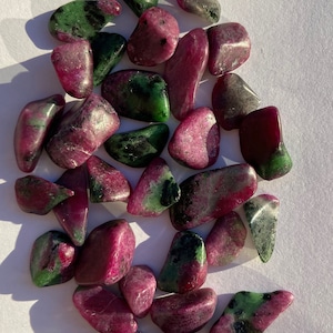 Könnte beinhalten: Eine Sammlung von grünen und rosafarbenen, getrommelten Edelsteinen. Die Steine sind glatt und poliert und haben verschiedene Formen und Größen.