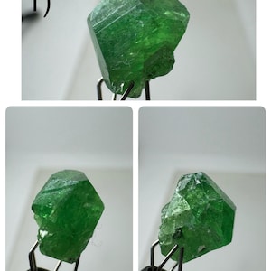 May include: A vibrant, emerald-green gemstone with a crystalline structure. The gem is held by metal tweezers, displaying its natural, irregular shape and vivid colour. The gem's facets reflect light, highlighting its clarity and depth.