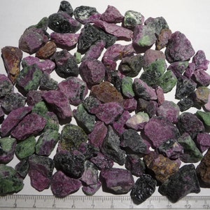 May include: A collection of rough, unpolished green and pink gemstones. The stones are various sizes and shapes, with some having a black, speckled appearance.