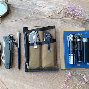 May include: A tan nylon pocket organizer with black trim containing a black pen and two folding knives with metal clips. Beside it are a black multi-tool, a silver pen, a black flashlight, a small silver flashlight, a black lighter, and a blue passport.