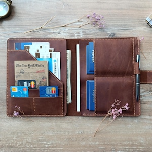 May include: Open brown leather wallet with multiple pockets filled with cards, cash, and documents. The wallet features a pen holder and a snap closure. A copy of The New York Times is visible in one of the pockets. The wallet is open on a wooden surface.