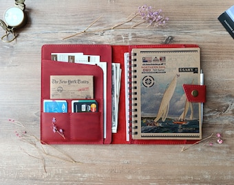 May include: Open red leather journal with a spiral-bound notebook featuring a vintage nautical illustration. The journal has pockets holding a newspaper, credit cards, and other items. A pen is secured by a red leather strap.