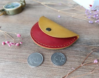 May include: A small, half-circle shaped leather coin purse with a red interior and a yellow exterior. The purse has a snap closure and is sitting on a wooden surface.