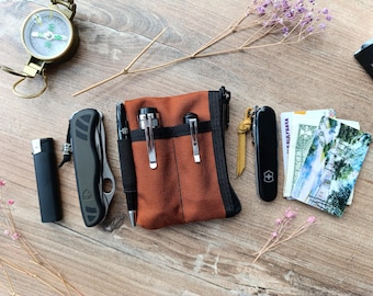 May include: A brown canvas pouch with a zipper closure, containing two pens, a lighter, a pocket knife, and a Swiss Army knife. The pouch is on a wooden surface with a compass and dried flowers.