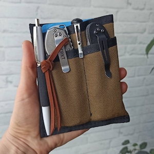 May include: A brown canvas utility pouch with multiple pockets, holding a pen, a small silver knife, a pair of tweezers, and a black folding knife. The pouch has a brown leather strap with the word "CRKT" embossed on it.