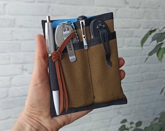 May include: A brown canvas utility pouch with multiple pockets, holding a pen, a small silver knife, a pair of tweezers, and a black folding knife. The pouch has a brown leather strap with the word "CRKT" embossed on it.