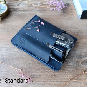 May include: A dark blue leather wallet with the name "JOHN" embossed on the front. The wallet has a pocket for a multi-tool and a pen. The multi-tool is a CRKT brand.