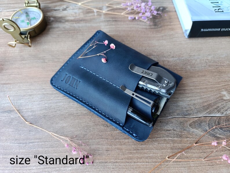 May include: A dark blue leather wallet with the name "JOHN" embossed on the front. The wallet has a pocket for a multi-tool and a pen. The multi-tool is a CRKT brand.