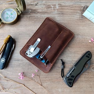 Könnte beinhalten: Eine braune Lederbörse mit einem Taschenmesser und einem Multitool im Inneren. Die Börse hat einen Klappverschluss und ist aus gealtertem Leder gefertigt.