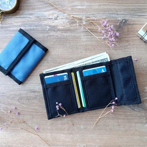 Könnte beinhalten: Ein blau-schwarzes Canvas-Portemonnaie, geöffnet, um Bargeld und Kreditkarten zu zeigen. Ein geschlossenes blaues Portemonnaie liegt daneben. Ein Kompass und ein Notizbuch liegen ebenfalls auf der Holzoberfläche. Ein praktisches Accessoire.