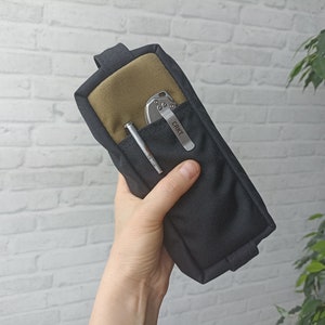 May include: A black and olive green pouch containing a pen and a silver folding knife with the text "CRKT" on the clip. The pouch is held in a hand against a white brick wall.