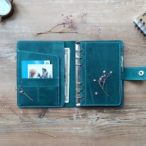 May include: A teal leather ring binder planner featuring a six-ring metal mechanism, a snap button closure, and interior pockets containing a one-dollar bill and a small photograph. The planner includes a pen loop and visible stitching.