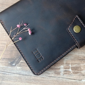 May include: A dark brown leather wallet with the word "FIDE" embossed on the front. The wallet has a snap closure with a brass button and is stitched with light brown thread. A sprig of dried flowers rests on the wallet.