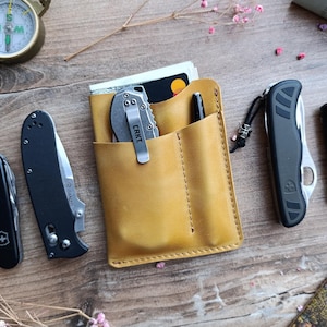 May include: A tan leather wallet with three pockets, a pen, and a small silver pocket knife. The knife has the word "CRKT" printed on it.