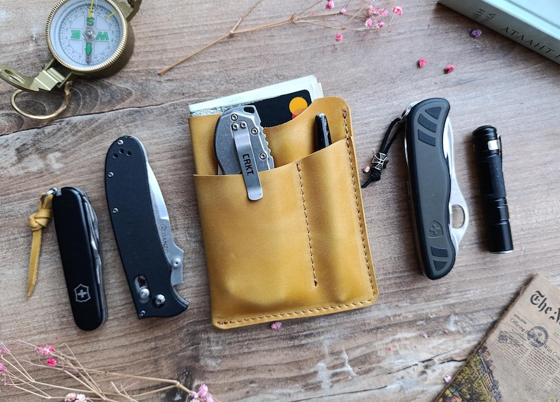 May include: A tan leather wallet with three pockets, a pen, and a small silver pocket knife. The knife has the word "CRKT" printed on it.