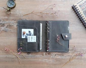 May include: A dark gray leather planner with a six-ring metal binder mechanism, a snap button closure, and internal pockets containing a photograph, a business card, and a one-dollar bill. A cat-shaped metal paperclip is attached.