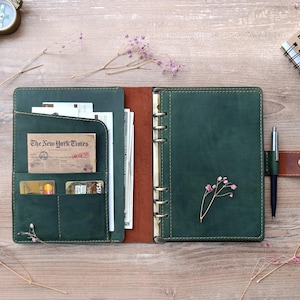 May include: A dark green leather planner with a six-ring metal binder mechanism. The interior features multiple card slots, a document pocket containing paper and currency, and a pen loop holding a silver pen with a brown leather strap.